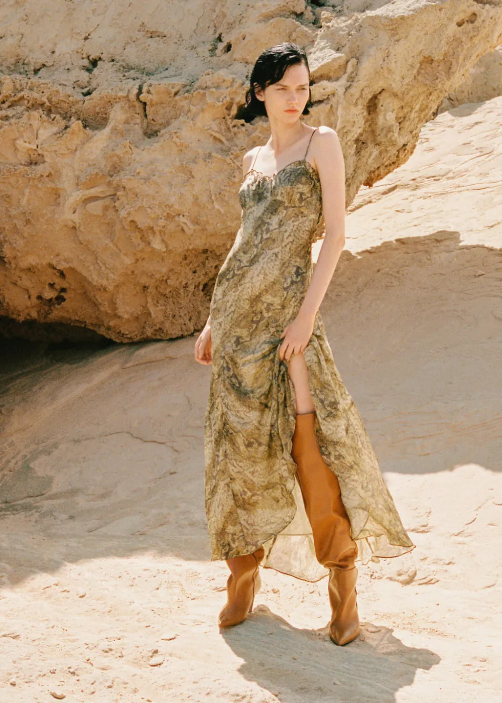 Elegant wedding guest outfits featuring a model in a flowing dress and stylish knee-high boots on a sandy backdrop.
